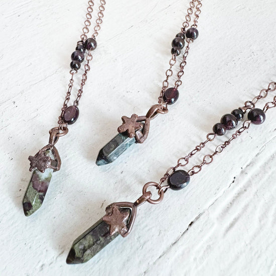 three copper electroformed gemstone spike necklaces with dark red beads and copper chain laid against a white wood background